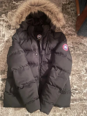 Svart dunjacka från Canada Goose - Qr kod fungerar. Svart pufferjacka från Canada Goose med stor fluffig huva och snygg pälsdetalj. Jackan har klassisk logga på ärmen, dragkedja och knappar framtill. Perfekt för kalla dagar och har en riktigt schysst oversized look.