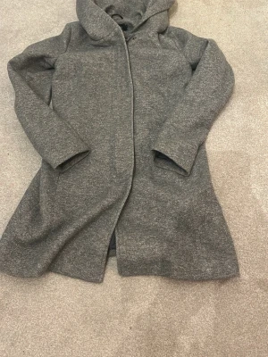 Kappa only  - Säljer en stilren grå kappa med huva och långa ärmar. Jackan har en rak passform och är tillverkad i ett mjukt material som känns skönt mot huden. 