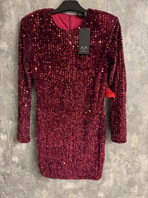 Vinröd paljettklänning från AX Paris - NY - Vinröd kort klänning från AX Paris, täckt med glittriga paljetter och sydd i mjuk sammet. Klänningen har långärmade ärmar och en rund halsringning. Perfekt för dig som vill sticka ut med en lyxig och glittrig look. Dragkedja bak för enkel påklädning.