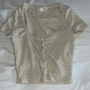 Beige kortärmad blus med knappar - Säljer en söt beige blus med korta ärmar och knappar framtill. Blusen har ett diskret mönster och en rundad halsringning. Perfekt för en avslappnad stil.