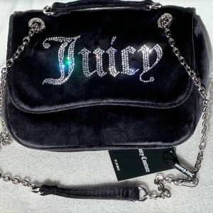 Svart axelväska Juicy Couture - * Axelväskan Kimberly från Juicy Couture i svart velour * Helt ny * Stilren och praktisk axelväska med en elegant klaffstängning * Har en justerbar rem som låter dig välja hur du vill bära den * Storlek i cm: 25x9x17 