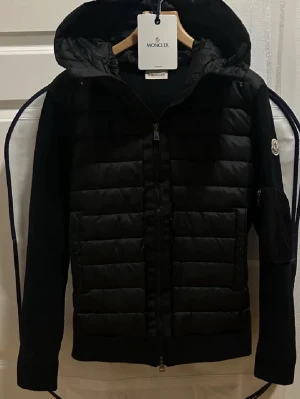 Svart pufferjacka från Moncler - Svart pufferjacka från Moncler med huva och ribbade stickade ärmar. Jackan har quiltad front, dragkedja och Moncler-logga på ärmen. Materialmix med polyester och stickad ull ger en schysst look. Perfekt för dig som vill ha både stil och värme.