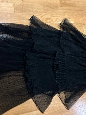 Svart prickig långkjol med volang 146/152 - Säljer en svart långkjol med tunna lager av transparent tyg och små prickar. I midjan är det en sammetsdetalj. Kjolen har flera volanger som ger ett snyggt och luftigt fall. Perfekt för dig som gillar en cool och trendig look med extra detaljer.