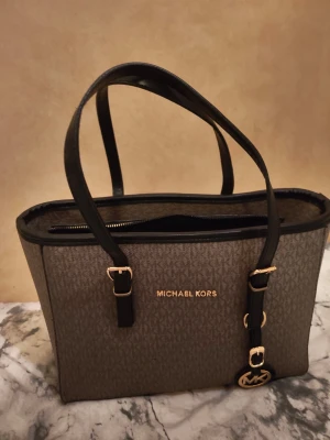 Brun handväska från Michael Kors - Snygg handväska från Michael Kors i brun färg med MK-mönster och svarta detaljer. Väskan har guldfärgade metalldetaljer, dragkedja upptill och flera fack inuti. Medföljer en justerbar axelrem i svart skinn. Perfekt storlek för allt du behöver ha med dig.