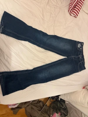 Victoria Beckham x Rock & Republic jeans - Mörkblå jeans med broderade kronor på bakfickorna från Victoria Beckham x Rock & Republic. Klassisk femficksmodell med raka ben och snygga kontrastsömmar. Tillverkade i slitstarkt denim med dragkedja och knapp framtill. storlek 27 och midjemått tvärsöver 36cm!