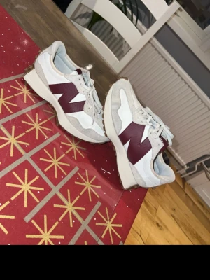 New Balance sneakers vit och vinröd 327 - Säljer ett par snygga New Balance sneakers i vit och vinröd färg med stor N-logga på sidan. Skorna har snörning, rund tå och en platt sula med gummidetaljer undertill. Ovandelen är i syntet och mocka, vilket ger en fräsch och sportig vibe.