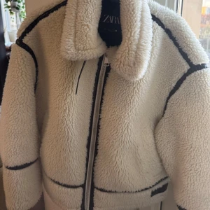 Vit teddyjacka från ZARA - Säljer en mysig vit teddyjacka från ZARA med svarta detaljer och dragkedja. Jackan har en hög krage och långa ärmar. Säljer då den inte kommer till användning, använd enstaka gånger, som ny. Storlek S!🤍