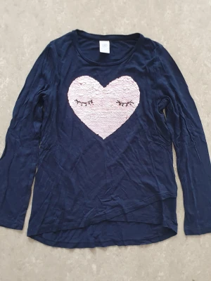 Tröja - Mörkblå långärmad tröja från Lindex med ett stort hjärta i rosa paljetter på bröstet. Hjärtat har vändbara paljetter som visar antingen ett sött ansikte med ögonfransar eller texten HELLO. Tröjan är i mjuk bomull och har rund hals.