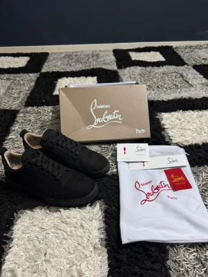 Svarta sneakers från Christian Louboutin - Säljer ett par svarta sneakers från Christian Louboutin med snyggt broderad logga på hälen och ikonisk röd sula. Skorna har snörning, rund tå och är tillverkade i mocka med skinnfoder. Perfekt för dig som vill ha en lyxig och stilren look.