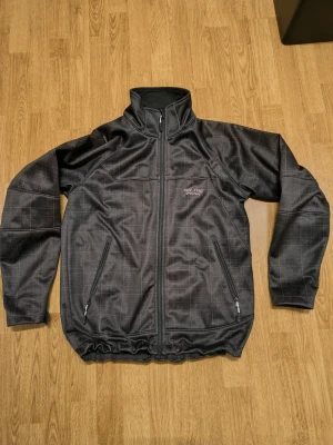 Svart vindjacka från On The Peak - Svart vindjacka med diskret rutigt mönster och hel dragkedja framtill. Jackan har två sidofickor med dragkedja, hög krage och långärmade ärmar.jackan har en sportig look med svensk flaggdetalj vid nederkanten.