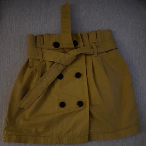 Gul kortkjol med bälte från Bershka - Säljer en snygg gul kortkjol från Bershka i storlek S. Kjolen har ett brett bälte i midjan och dekorativa svarta knappar framtill. Modellen är A-linjeformad och tillverkad i bomull, vilket gör den både bekväm och lätt att matcha med olika toppar.