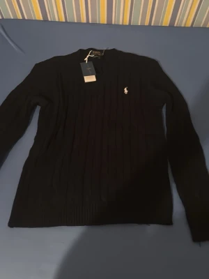 Svart stickad tröja från Polo Ralph Lauren - Klassisk svart stickad tröja från Polo Ralph Lauren med diskret vit logga på bröstet. Tröjan har lång ärm, ribbade muddar och ett stilrent, smalt snitt. Perfekt till lager på lager och enkel att matcha med allt.helt ny storlek m 