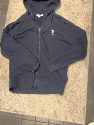 Marinblå hoodie från USPA - Säljer en marinblå hoodie från USPA med dragkedja och huva. Tröjan har långa ärmar och ett litet broderat logomärke på bröstet. Perfekt för chill dagar och enkel att matcha med jeans eller joggers.