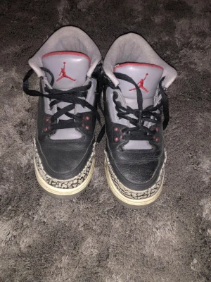 Nike Air Jordan 3 Retro Black Cement - Nike Air Jordan 3 Retro Black Cement med svart och grått skinn, röda detaljer och ikoniskt elefantmönster på tå och häl. Skorna har snörning, rund tå och platt sula. Klassisk Jumpman-logga på plösen och AIR-tryck på hälen. Perfekt för dig som gillar streetwear och sneakers. 