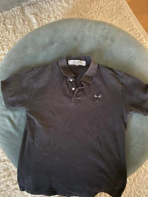 Svart Comme des Garçons PLAY piké - Säljer en svart piké från Comme des Garçons PLAY med klassisk krage och två knappar. På bröstet sitter det ikoniska hjärtemärket med ögon i svart. Skön passform och stilren look som funkar till allt. Tröjan är i storlek L men passar M väldigt bra. Nypris 1400kr 