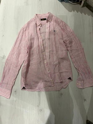 Randig linneskjorta från Morris - Säljer en snygg rosa och vit randig skjorta från Morris i storlek L. Skjortan är tillverkad i luftig linne, har klassisk krage, långa ärmar och knappar framtill. Broderad logga på bröstet och diskret etikett vid nederkanten. Perfekt för en chill och stilren look.