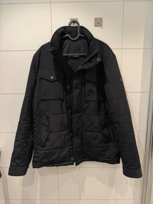 J.Lindeberg jacka i strl L - Snygg och stilren jacka från J.Lindberg med hög krage, långa ärmar och flera fickor med både knappar och dragkedjor. Jackan har quiltad design och stängs med både knappar och dragkedja. Perfekt för kalla dagar och har en clean, modern look.