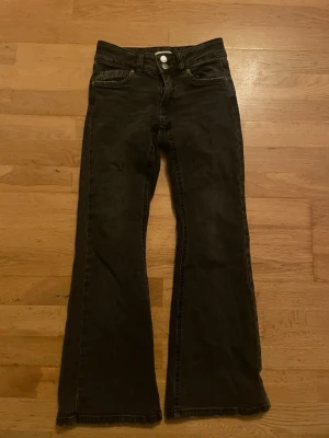 Svarta bootcut jeans från Denim Day - Säljer ett par svarta bootcut jeans från Denim Day i storlek 146. Jeansen har hög midja, två knappar fram och snygga fickor med knapp bak. Materialet är klassisk jeans och passformen är loose med breda ben. Perfekt för dig som gillar retrovibbar och cool stil.