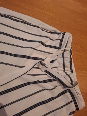 Randiga wide byxor från Black Stormy - Snygga vita byxor med svarta vertikala ränder från Black Stormy. Modellen är wide och har en midwaist passform med knytband i midjan. Materialet känns mjukt och luftigt, perfekt för dig som gillar en chill och stilren look.