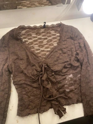 Brun spetsblus med snörning - Säljer en brun långärmad blus i spets med blommigt mönster. Toppen har v-ringning, snörning framtill och fina volanger.Perfekt  att styla med jeans eller kjol.