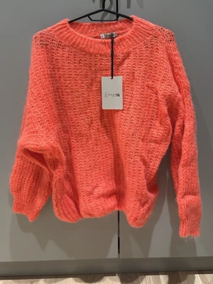 Stickad korallrosa tröja från DesignSi - Supermjuk och fluffig stickad tröja i en härlig korallrosa färg från DesignSi. Tröjan har en loose passform, bred ribbad mudd vid ärmslut och nederkant samt lång ärm. Perfekt för lager-på-lager under kyliga dagar.