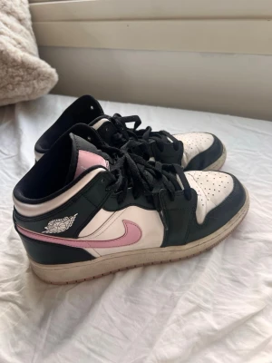 Nike Air Jordan 1 svart/vit/rosa - Säljer ett par Nike Air Jordan 1 sneakers i svart, vitt och rosa. Skorna har klassisk snörning, svart läderpanel, vit tåbox och rosa swoosh samt detaljer på tungan. Perfekta för dig som gillar streetstyle och vill sticka ut med färg.