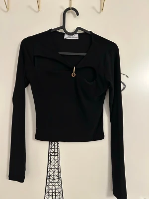 Svart långärmad topp med cut-out från Chiquelle - Svart långärmad topp från Chiquelle i storlek XS. Toppen har en cut-out detalj framtill med en guldfärgad ring och är croppad i modellen. Observera och se 3:e bilden att toppen är nopprig. Detta är inget som påverkar dess funktion och är synligt vid nära håll och vid inspektion. 