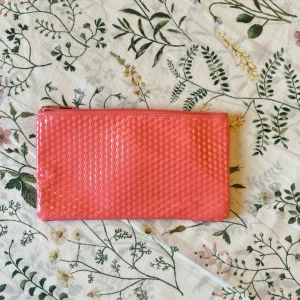 Rosa kuvertväska/necessär med mönstrad yta - Snygg rosa kuvertväska/necessär med glansig, geometriskt mönstrad yta och dragkedja upptill. Perfekt storlek för småsaker och enkel att ta med sig. Väskan är gjord i syntetmaterial och har ett stilrent, modernt uttryck.