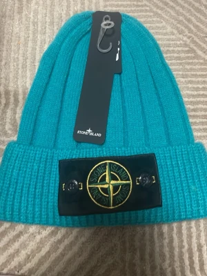 Turkos stickad mössa från Stone Island - Säljer en turkos stickad mössa från Stone Island med den klassiska logotypen framtill på en svart patch. Mössan har ribbad struktur och är riktigt mjuk och skön. Perfekt för att hålla värmen och samtidigt se snygg ut.