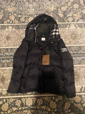Svart dunjacka från Burberry - Säljer en svart dunjacka från Burberry med avtagbar huva och klassiskt rutigt foder. Jackan har dragkedja, knappar och två stora fickor framtill. På ärmen finns en vit Burberry-logga. Perfekt för kalla dagar och riktigt snygg streetstil.