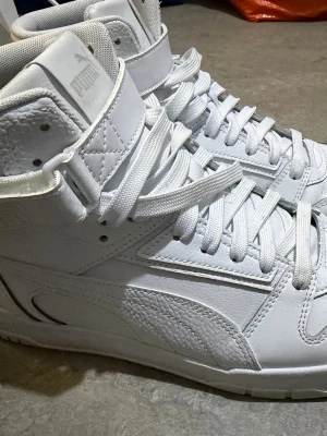 Vita höga sneakers från Puma strl 44 - Säljer ett par fräscha vita höga sneakers från Puma i storlek 44. Skorna har snörning, rund tå och platt sula. Tillverkade i skinn med detaljerad sömnad och en bred rem över vristen. Perfekta för dig som gillar klassisk streetstil.