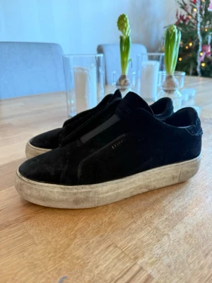 Svarta Arigato sneakers i mocka - Svarta slip-on sneakers från Arigato med ovandel i mjuk mocka och elastiskt band över foten. Skorna har en vit platt gummisula med Arigato-logga undertill och rund tå. Perfekt för dig som gillar minimalistisk stil och vill ha något som funkar till jeans eller shorts.