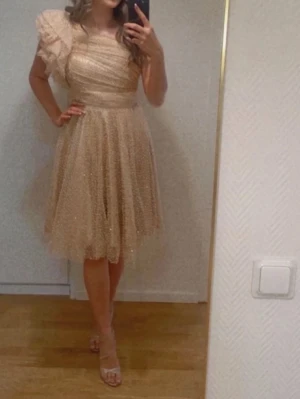 Champagnefärgad prickig omlottklänning - Säljer en champagnefärgad kortklänning med ett prickigt mönster och omlott-design. Klänningen har en axel med volang och är oneshoulder, vilket ger en unik och trendig look. Materialet är lätt och ger ett glittrigt intryck, perfekt för att sticka ut. Passformen är normal och kjolen är luftig.