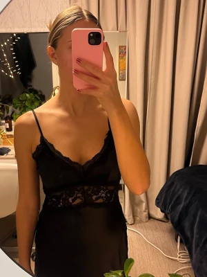 Svart långklänning med spetsdetaljer - Svart långklänning i satin med spetsdetaljer och smala axelband. Klänningen har en slits och öppen rygg. Från Gina Tricot. Oanvänd med prislapp kvar. Skickas eller kan hämtas i Partille.