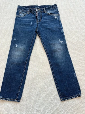 Blå jeans från Dsquared2 - Säljer ett par blå jeans från Dsquared2 med klassisk femficksdesign och snygga slitningar. Jeansen har raka ben, normal passform och detaljerade sömmar med kontrastfärg. Materialet är robust denim i bomull och de har en läderpatch bak i midjan.