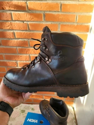 Bruna kängor från Alico i skinn 43 - Säljer ett par robusta kängor från Alico, tillverkade i mörkbrunt skinn med grov sula och snörning med metallöglor. Perfekta för dig som gillar äventyr och vill ha slitstarka boots med klassisk outdoor-look. Fodrade och med rund tå för extra komfort. Tillverkade I Italien 