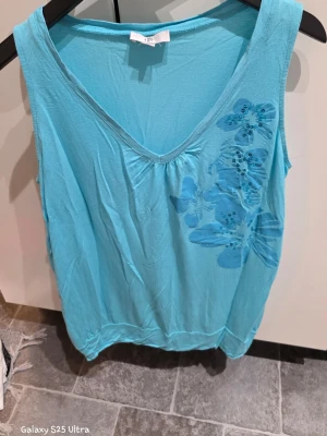 Blå ärmlös linnetopp med blommor - Snygg ljusblå linnetopp från H&M i storlek 44/46. Toppen har v-ringning och är ärmlös med ett stort blommigt tryck och paljettdetaljer på framsidan. Perfekt för varma dagar och enkel att matcha med jeans eller shorts.