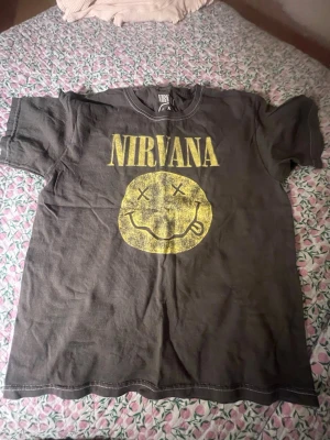 Svart Nirvana t-shirt med tryck - Cool svart t-shirt från Nirvana med ikoniskt gult smiley-tryck och stor logga på bröstet. Klassisk rund hals och korta ärmar. Perfekt för dig som gillar bandmerch och gillar lite oversized men inte för mycket! Denna är köpt i USA så vet inte exakt storlek, men har passat alla kring denna ålder. Trycket är meningen att den ska vara lite sliten så inget konstigt om du tycker den ser sliten ut! Tack ❤️