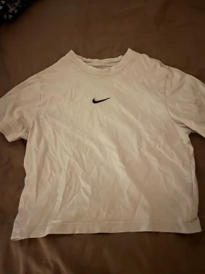 Vit Nike t-shirt med logga - Klassisk vit t-shirt från Nike med svart swoosh-logga på bröstet. Enkel och clean design med rund hals och korta ärmar. Tillverkad i mjuk bomull som känns skön mot huden. Perfekt till alla tillfällen där du vill ha en stilren look.