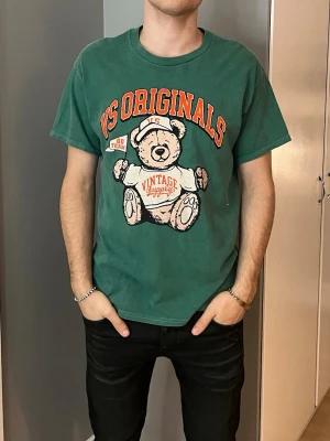 Grön VS Originals t-shirt med björn - Grön t-shirt från Vintage Supply med stor björn- och textprint i orange och vitt på bröstet. Klassisk rund hals och korta ärmar. Skön passform och mjukt material som känns chill och avslappnat.