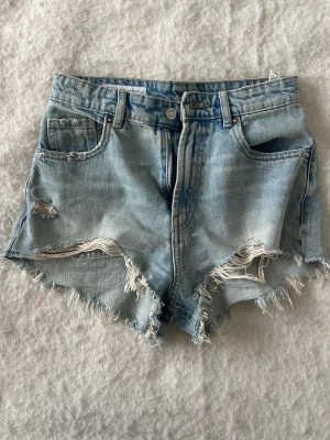 Ljusa denimshorts från Zara - Säljer ett par ljusblå denimshorts från Zara i storlek 34. De har fransiga kanter, slitna detaljer och klassisk femficksdesign. Shortsen är midwaist och har dragkedja och knapp framtill. Perfekta för en chill sommardag.