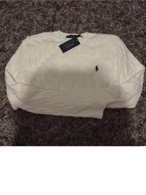 Vit stickad tröja från Polo Ralph Lauren - Säljer en vit stickad tröja från Polo Ralph Lauren med klassisk kabelstickning och rund hals. Tröjan har långärmade och ett broderat marinblått Polo-logo på bröstet. Perfekt för dig som gillar stilrena och tidlösa plagg.