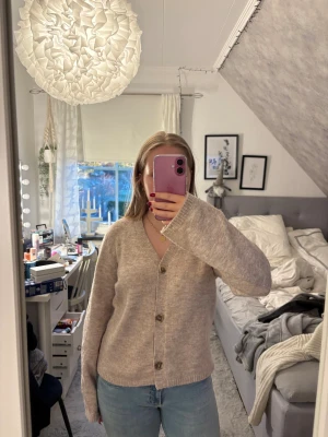 Beige stickad kofta med knappar - Mysig beige stickad kofta med v-ringning och stora knappar framtill. Koftan har lång ärm och en avslappnad passform, perfekt att slänga på sig över en t-shirt eller linne. Enkel och stilren design som funkar till det mesta.