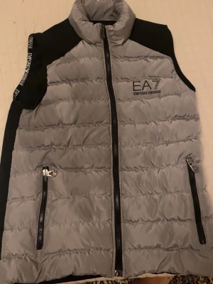 Grå dunväst från Emporio Armani - Säljer en stilren grå dunväst från Emporio Armani EA7 med svarta detaljer. Västen har hög krage, två fickor med dragkedja och logga på bröstet. Svarta partier på axlar och rygg samt tydliga quiltade sömmar framtill. Perfekt för lager på lager och streetwear.