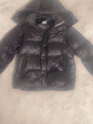 Svart pufferjacka från Burberry - Svart pufferjacka från Burberry med huva och quiltad design. Jackan har en snygg Burberry-logga på ärmen och dragkedja framtill. Insidan har ett klassiskt rutigt Burberry-mönster i grå toner. Perfekt för kalla dagar och riktigt stilren. Går även att ta av armarna för att göra jackan till en väst vilket gör den perfekt för våren.