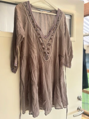 Beige klänning med broderi - Säljer en beige kortklänning med v-ringad hals och broderade detaljer i lila och brunt. Klänningen har trekvartsärm och är tillverkad i ett lätt bomullstyg. 