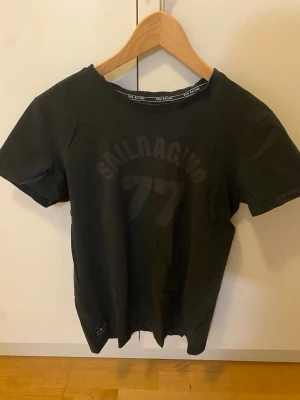 Svart t-shirt från Sail Racing - Svart t-shirt från Sail Racing med tryck 'SAILRACING 77' på bröstet och diskret logga nertill på framsidan. Strl S.
