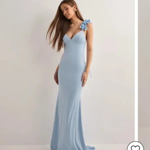 Ljusblå långklänning med blomdetalj - Supersnygg ljusblå långklänning med figurnära passform och v-ringning. Klänningen har en axelbandslös design på ena sidan och en axel med fina blommor som detalj. Perfekt för bal eller festliga tillfällen. (Använd en gång och nypris är 1300)