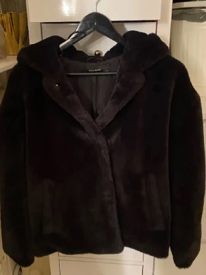 Svart pälsjacka med huva från Zara - Mjuk och fluffig svart pälsjacka från Zara med stor huva och diskreta fickor. Jackan har en enkel, clean look och är perfekt för dig som gillar att sticka ut med en cozy vibe. Passformen är normal och jackan är helsvart utan mönster.