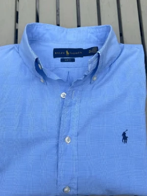 Blå slim fit skjorta från Ralph Lauren - Snygg blå skjorta från Ralph Lauren i slim fit-modell med diskret rutigt mönster. Klassisk button-down krage, knappar framtill och ikonisk broderad logga på bröstet. Perfekt för dig som gillar stilrena och fräscha plagg. Tror storlek M